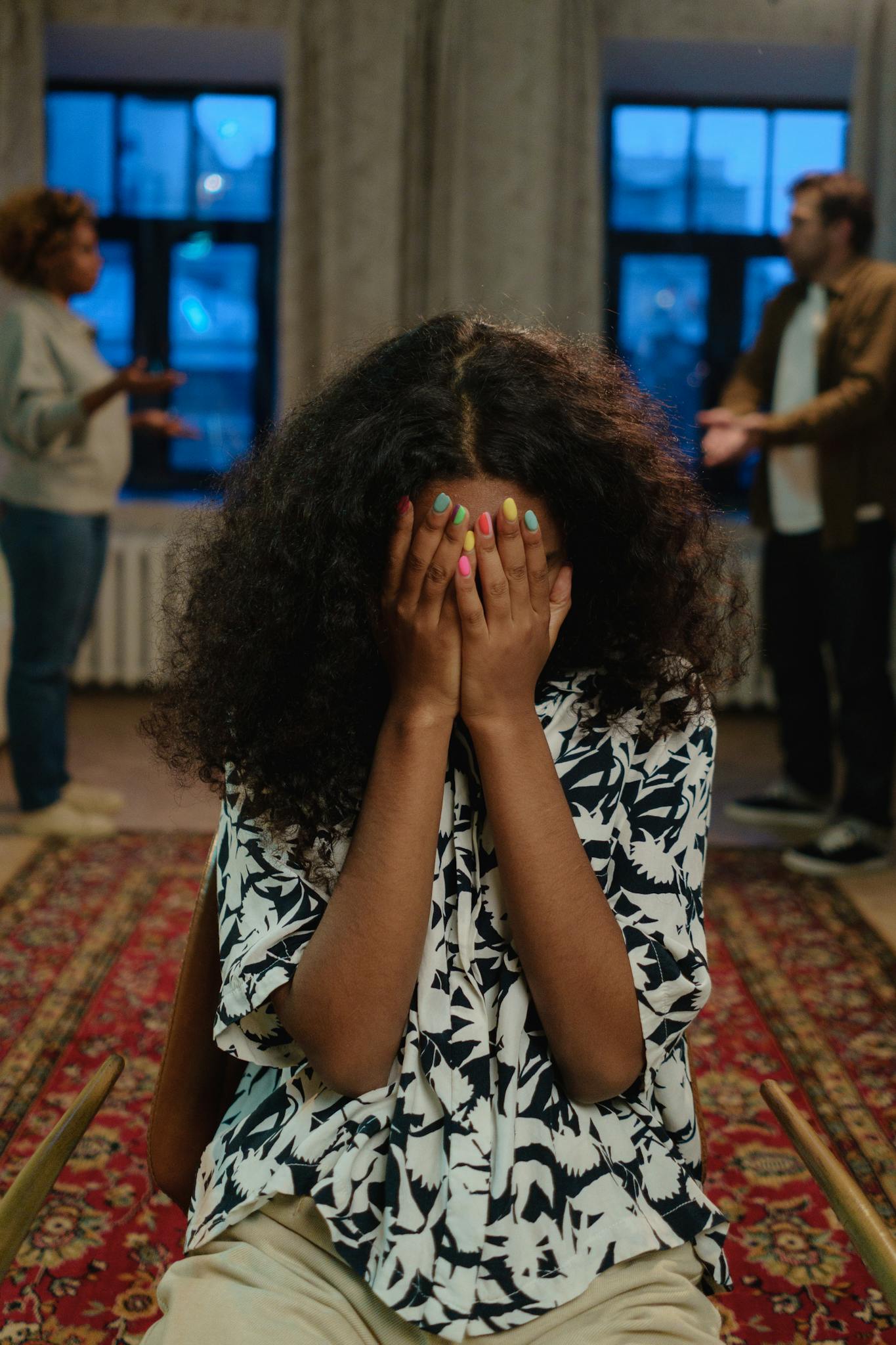 A young woman sits indoors with her face covered amidst a family argument, conveying distress and conflict.